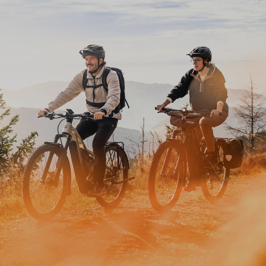 E-MTB Trail Pärchen auf Fahrrädern fährt durch eine Wolke aus rotem Staub im Sonnenlicht.