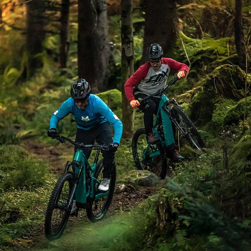 Zwei Mountainbiker fahren mit E-MTBs sportlich durch einen moosbedeckten Waldtrail.