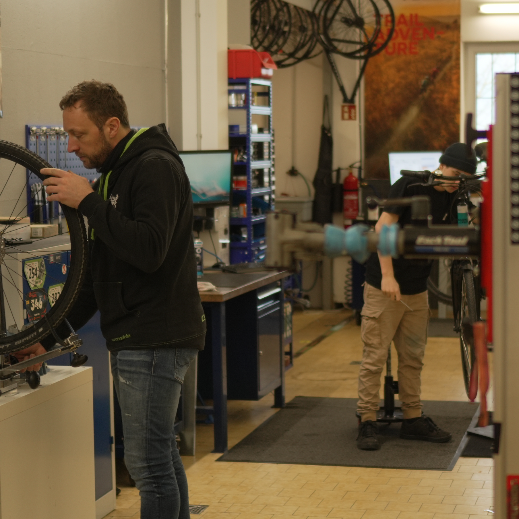 Zwei Fahrradmechaniker bei der Arbeit in der Werkstatt – einer prüft ein Laufrad, der andere schraubt an einem Bike auf dem Montageständer.