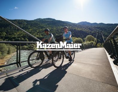 Kazenmaier Leasing Logo auf Hintergrund mit zwei Radfahrern die ĂĽber eine BrĂĽcke fahren